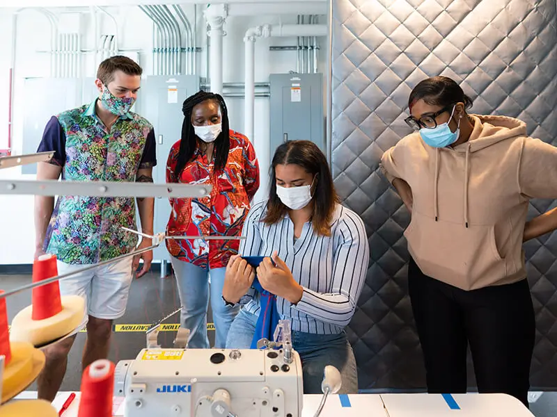 Um grupo de pessoas observando uma mulher trabalhando em uma máquina JUKI