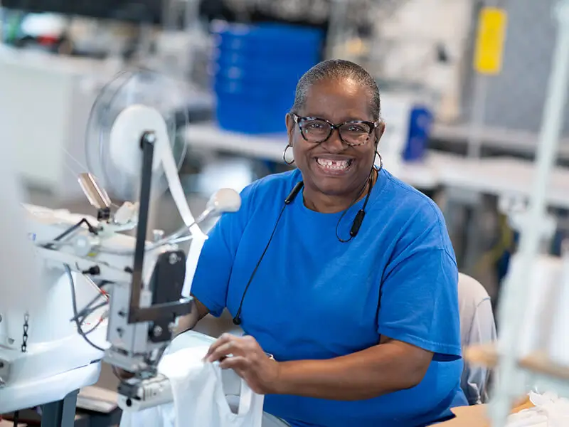 Uma mulher sorrindo para a câmera, ela está ao lado de uma máquina JUKI